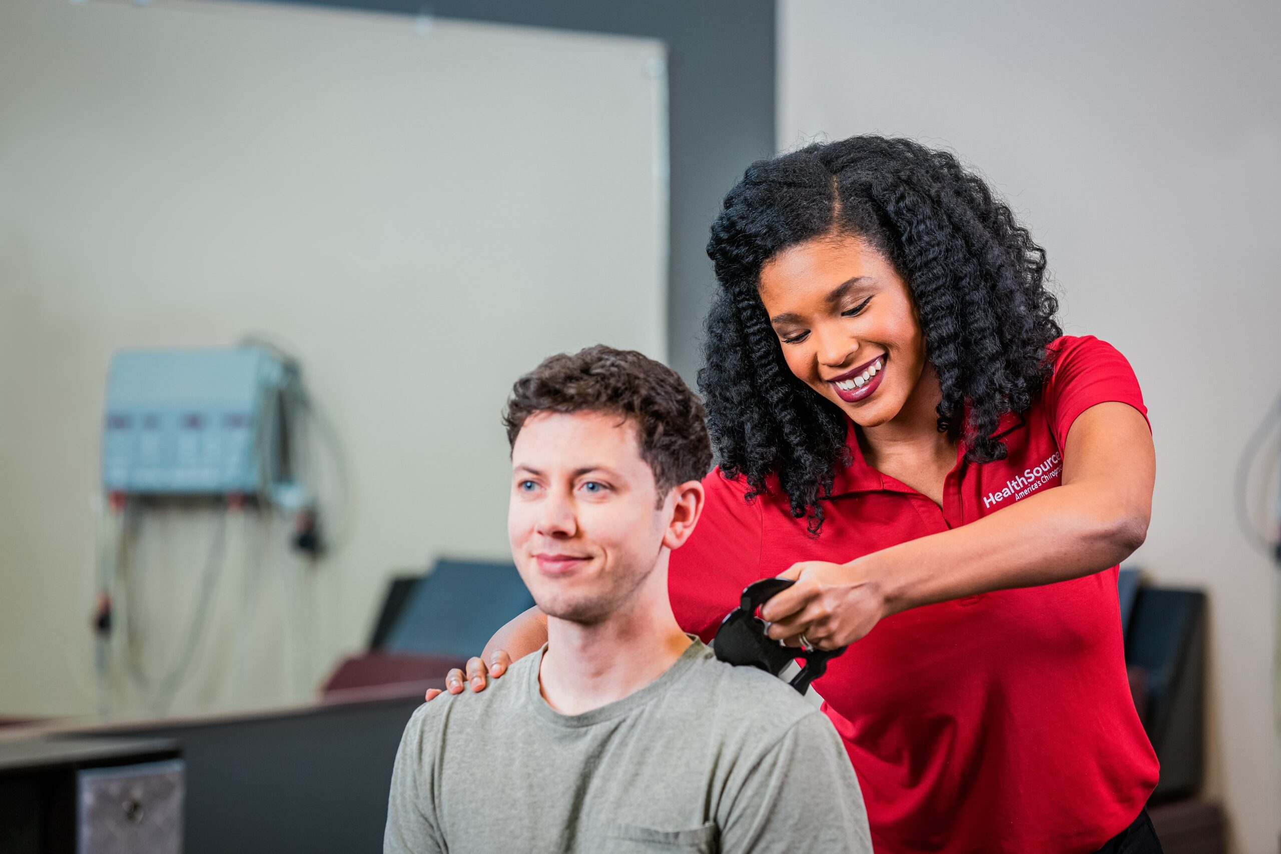 Patient sits facing the camera as a practitioner massages their shoulders using a specialty tool.