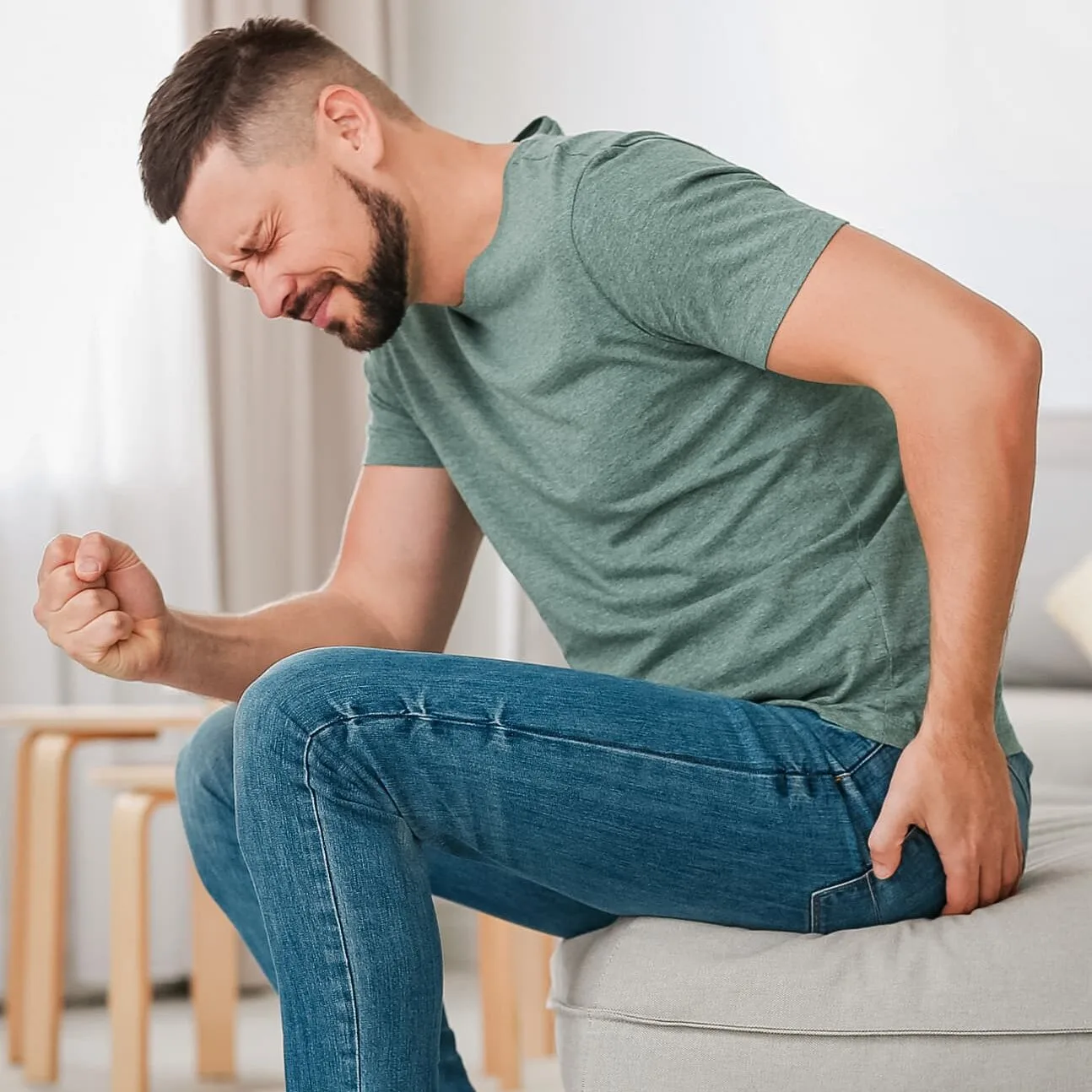 A man sits on the edge of his couch and clutches his thigh in pain.